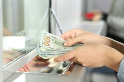 Our customers include currency exchange offices and financial service providers A close-up view of a person’s hands holding US $100 bills at a currency exchange counter, with a protective glass barrier in between